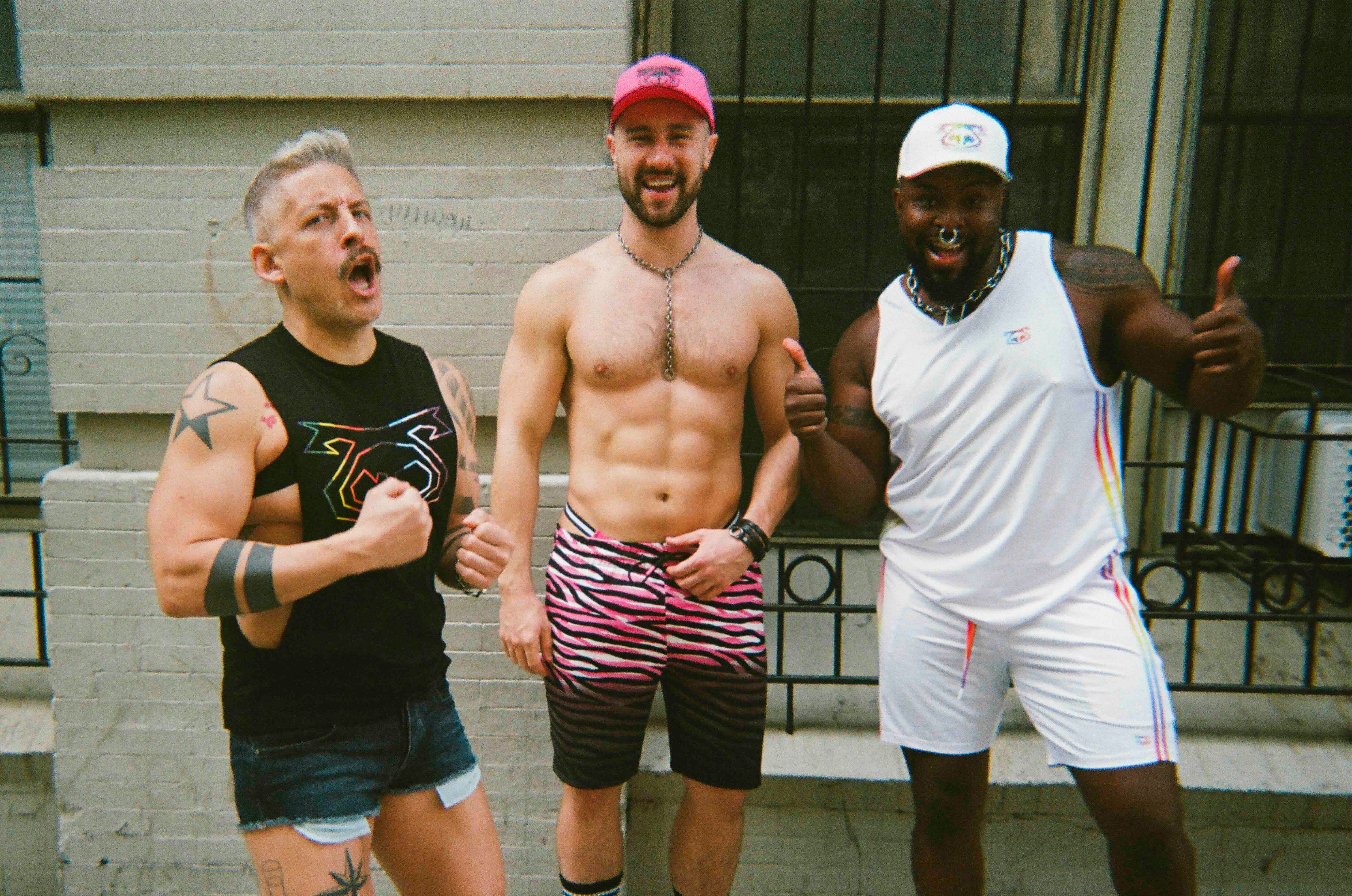 Three men posing outdoors, two of whom are shirtless, with a building in the background.